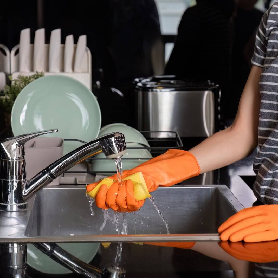 Person using orange gloves to wash dishes with a sponge in a modern kitchen sink.