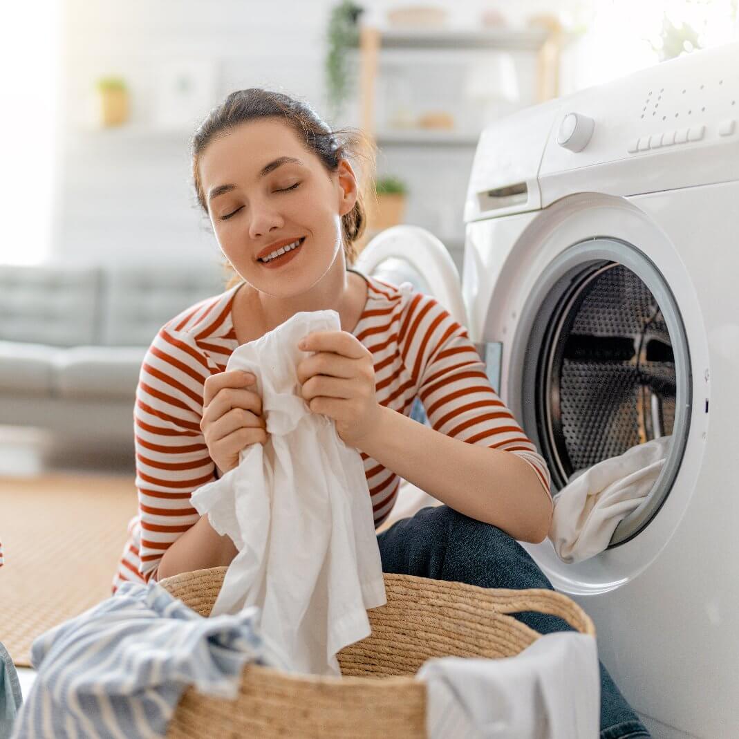 Woman enjoying fresh laundry scent while doing laundry at home in a bright, cozy setting.