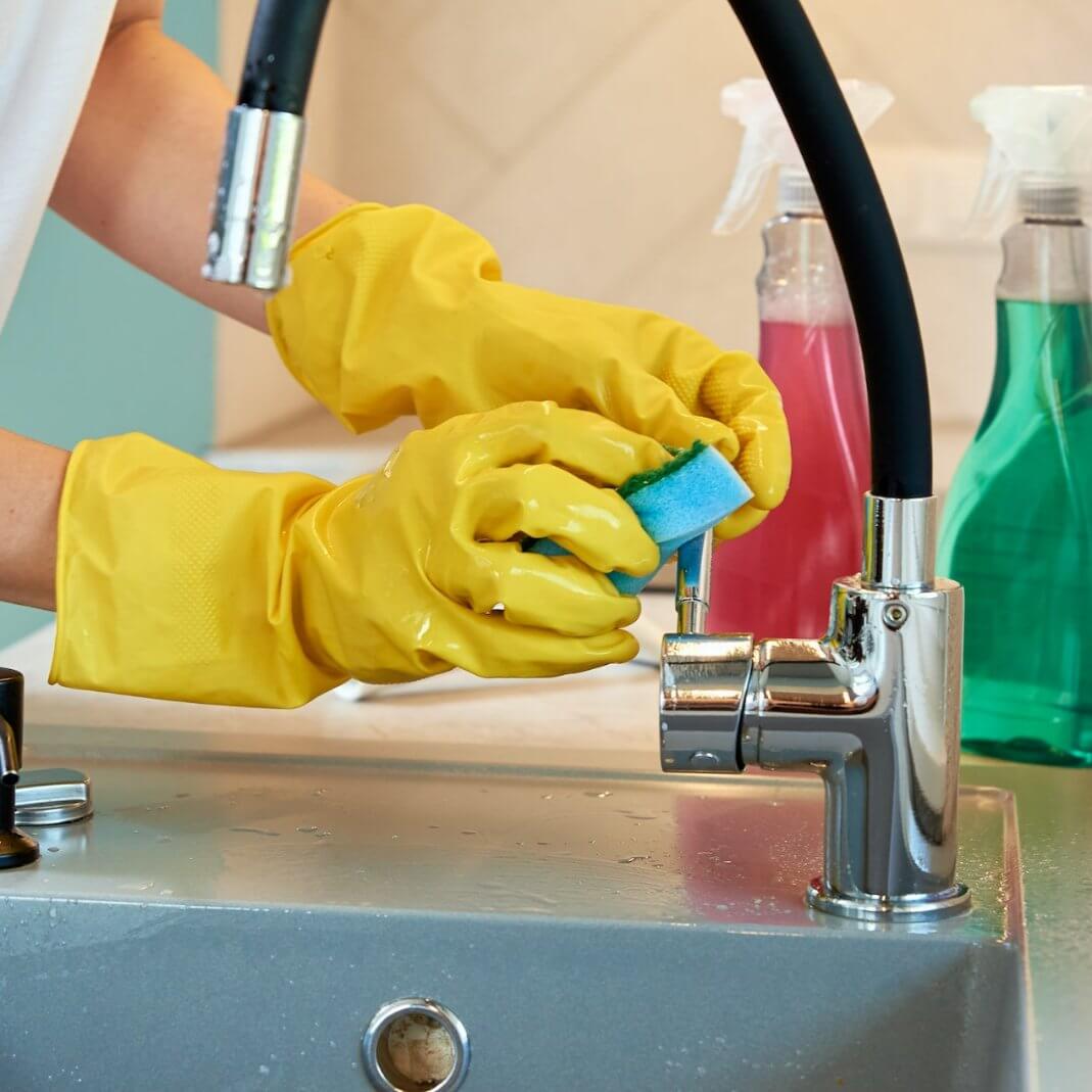 Person in yellow gloves scrubbing a sink with cleaners in colorful bottles nearby.